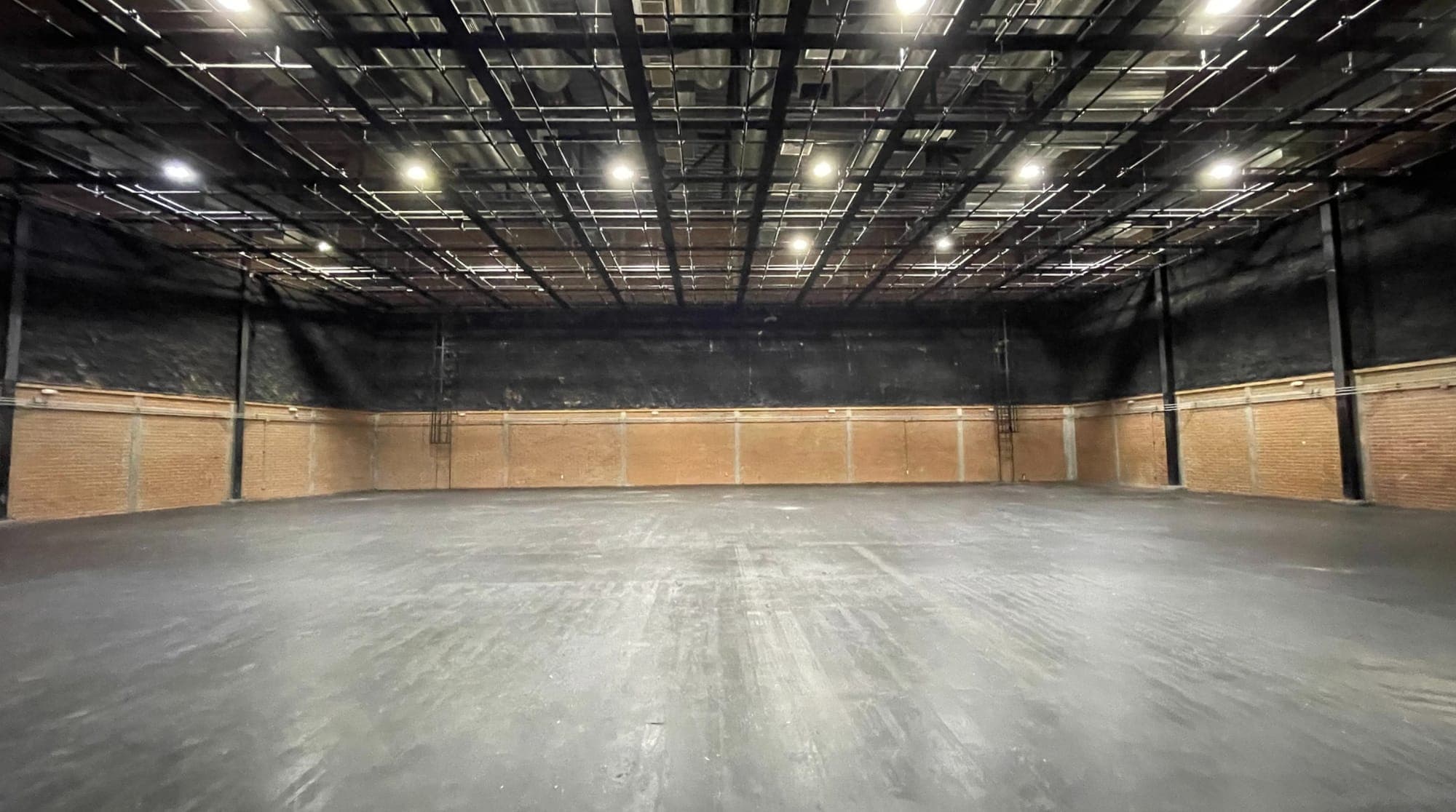Nave vacía vista desde el acceso: piso de concreto, muros negros con zócalo de ladrillo y la parrilla de iluminación cubriendo todo el techo.