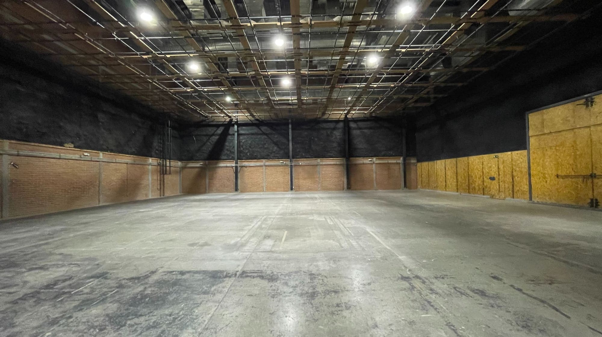 Nave vacía de fondo profundo: piso de concreto, muros negros sobre zócalo de ladrillo con columnas metálicas, un costado forrado de tablero de madera con portón y la parrilla de iluminación encendida en todo el techo.