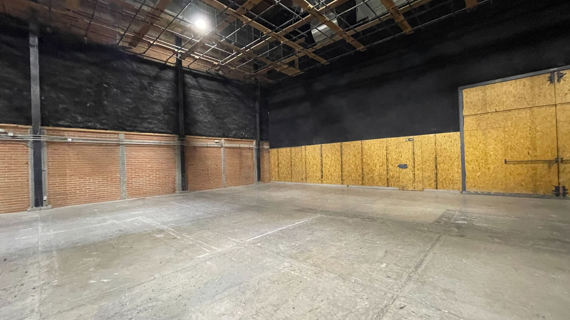 Nave vacía con piso de concreto: muros negros sobre zócalo de ladrillo, un frente forrado de tablero de madera con puerta y portón, y la parrilla de iluminación en el techo.