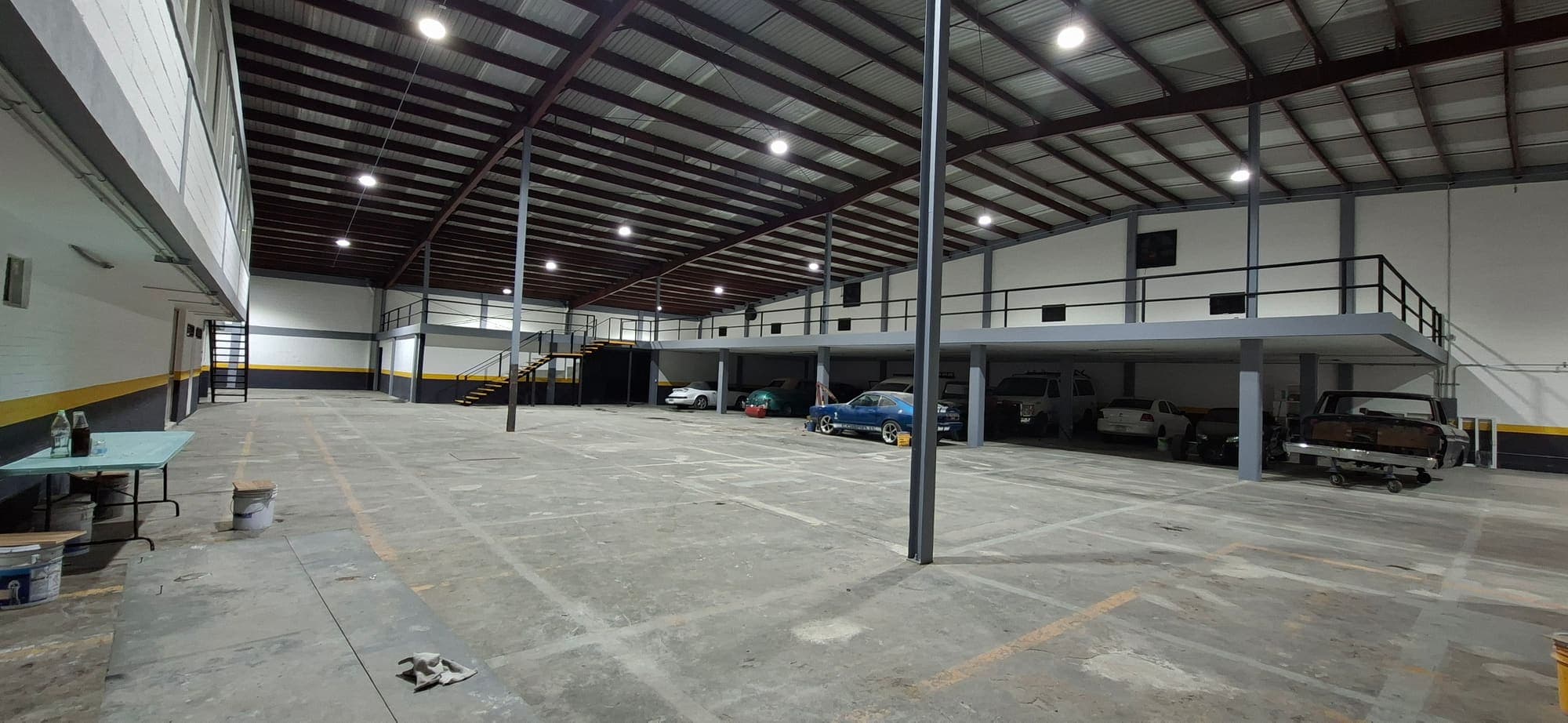 Nave de techo alto con estructura metálica y luminarias de campana; al fondo, un mezzanine con barandal y vehículos guardados debajo.