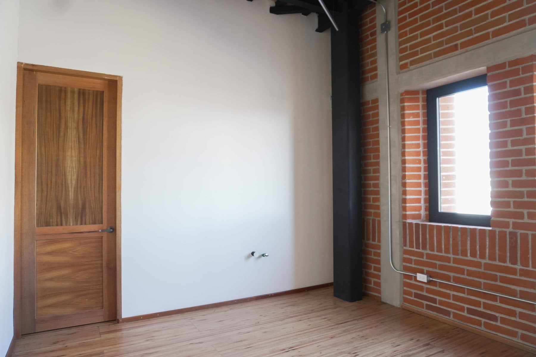 Camerino con puerta de madera y ventana enfrentadas, y columna metálica negra al centro.