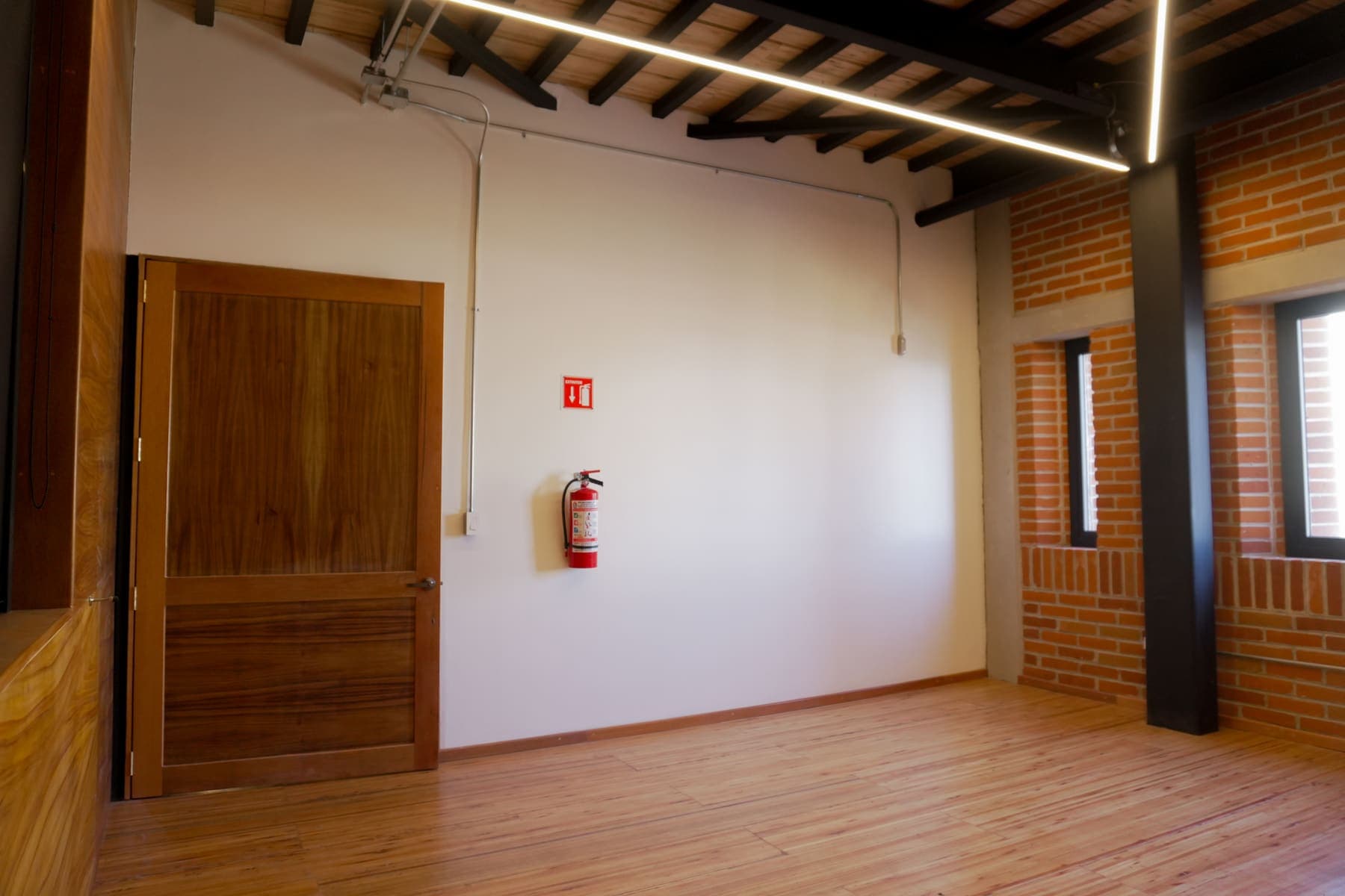 Camerino vacío de piso de duela, muro blanco y ladrillo visto, con puerta de madera y luminaria lineal.