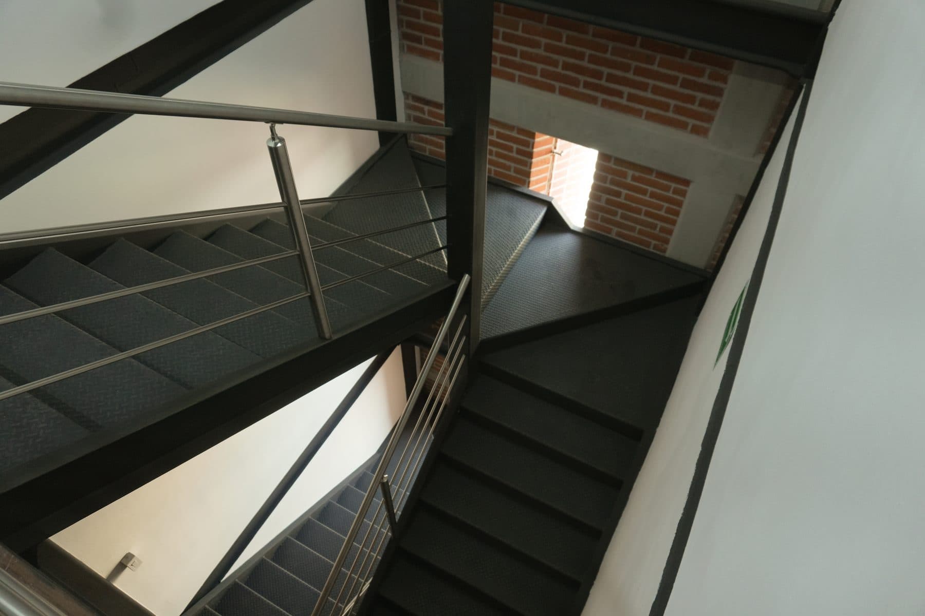 Escalera de peldaños metálicos y barandal de tubo, entre muro blanco y muro de ladrillo.