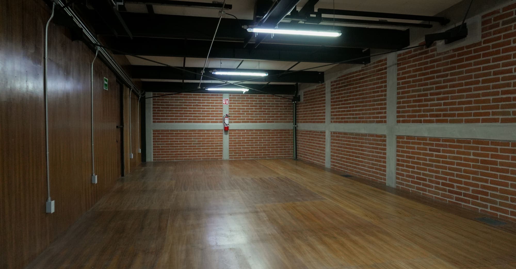 Planta diáfana de piso de duela, con muro de ladrillo, columnas encaladas y luminarias suspendidas del techo.