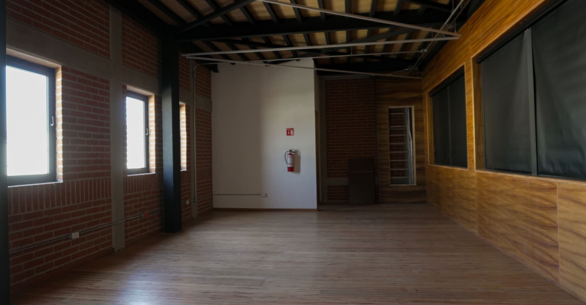 Sala de techo de viga vista con ventanas altas a un costado y persianas oscuras al otro.
